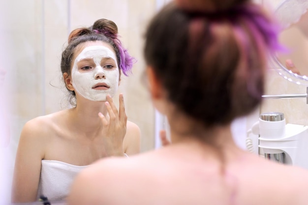 Mujer hermosa joven con máscara de curación natural cosmética blanca en la cara en el baño. Cosmetología, hidratación y facial en la adolescencia, piel limpia y perfecta