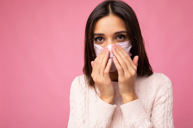 Mujer hermosa joven con máscara protectora de virus reutilizable en la cara contra el coronavirus y temeroso de aislado en la pared de fondo rosa.