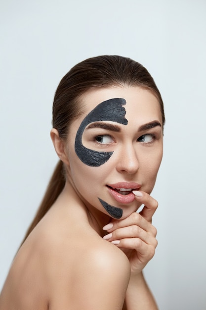 Mujer hermosa joven en una máscara para el rostro de la arcilla negra terapéutica. Cuidado de la piel de la cara de belleza. Tratamiento de spa . Modelo de niña con mascarilla cosmética hidratante.