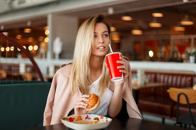 Mujer hermosa joven modelo en una chaqueta de moda rosa comiendo comida rápida en un café, bebe una cola de una taza roja