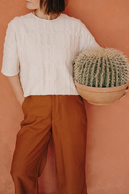 Mujer hermosa joven en pantalones de jengibre y camiseta blanca sosteniendo cactus en una olla contra la pared de jengibre