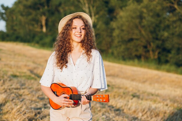 Mujer hermosa joven con el pelo largo y rizado en pantalones cortos, un sombrero con una guitarra posa en un campo de trigo en el verano al atardecer