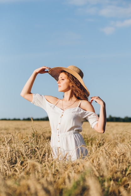 Mujer hermosa joven con el pelo largo y rizado posa en un campo de trigo en el verano al atardecer