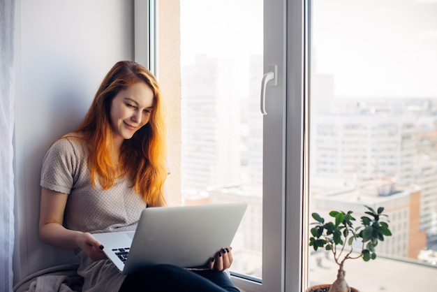 Mujer hermosa joven con pelo largo rojo se sienta en el alféizar de la ventana y mira la computadora portátil. Escritora con pc en su regazo en el fondo de la ventana en el apartamento de la ciudad. Aprendizaje en línea, concepto autónomo.