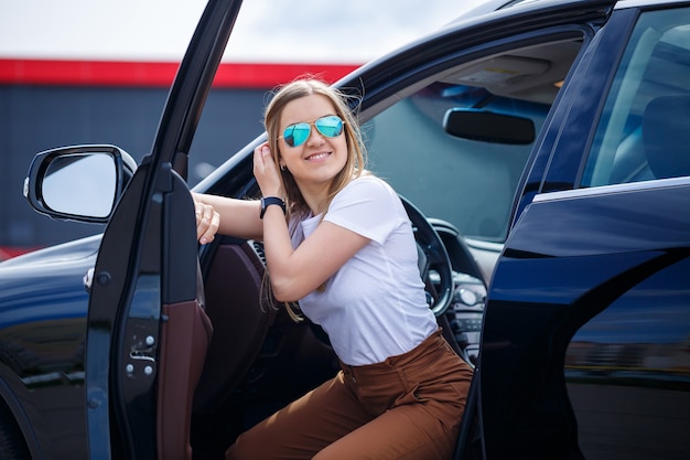 Mujer hermosa joven con el pelo largo se sienta en un coche negro en un estacionamiento. Chica guapa en ropa casual. Viaje en coche