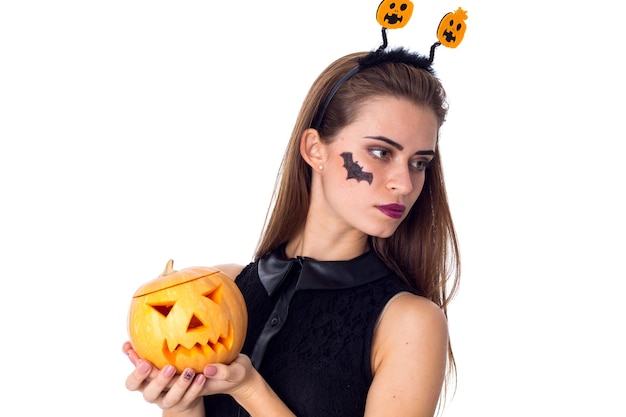 Mujer hermosa joven con una pequeña foto de murciélago en su mejilla en vestido negro sosteniendo una calabaza