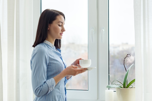 Mujer hermosa joven de pie en casa cerca de la ventana con una taza de café, espacio de temporada de invierno, cortinas de luz, espacio de copia