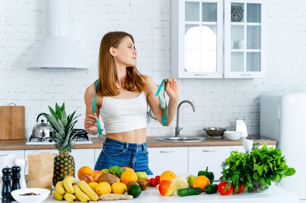 Mujer hermosa joven de pie cerca de la mesa llena de diferentes frutas y verduras Mujer sosteniendo cinta métrica Cocina ligera moderna