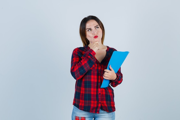 Mujer hermosa joven de pie en pose de pensamiento, sosteniendo la carpeta en camisa casual, jeans y mirando desconcertado. vista frontal.