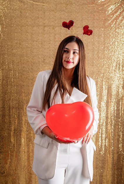 Mujer hermosa joven de pie sobre un fondo dorado brillante sosteniendo globos de corazón rojo