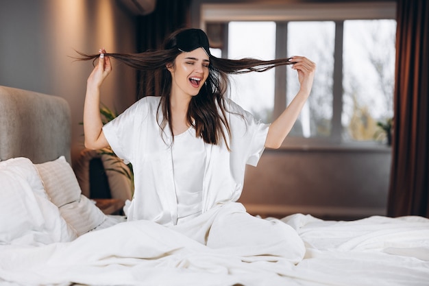 Mujer hermosa joven en pijama de seda blanca se despierta en el dormitorio en la cama. Una mujer se despierta de buen humor y sostiene su cabello en sus manos. Por la mañana o por la noche en el dormitorio con una modelo femenina.