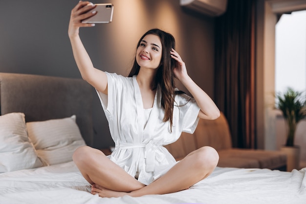 Mujer hermosa joven en pijama de seda blanca hace un selfie en el teléfono. Una modelo femenina con un teléfono en sus manos se sienta en la cama. por la mañana o por la noche en el dormitorio con una modelo femenina.