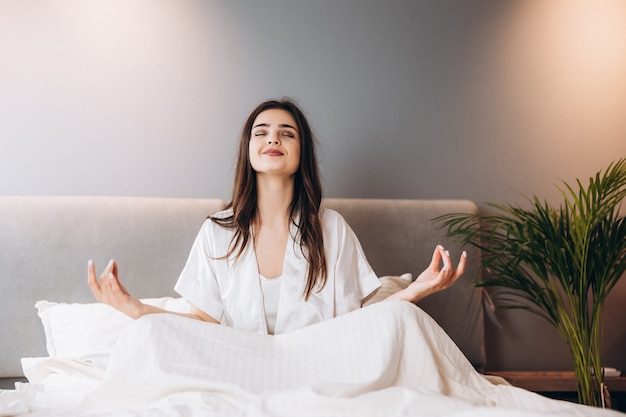 Foto mujer hermosa joven en pijama de seda blanco está haciendo yoga en el dormitorio en la cama. modelo femenino en posición de loto en la cama. por la mañana o por la noche en el dormitorio con una modelo femenina.