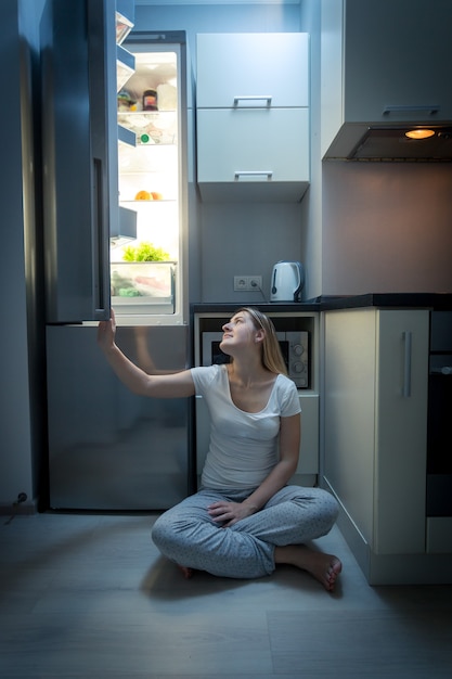 Mujer hermosa joven en pijama sentada en el piso de la cocina y mirando el refrigerador abierto en la noche