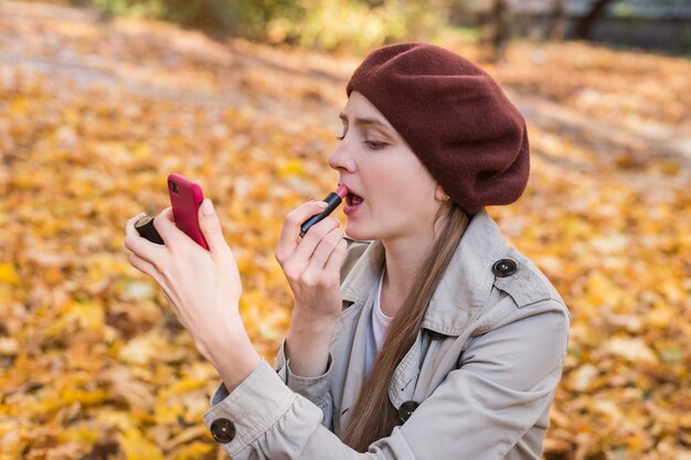 Mujer hermosa joven poner lápiz labial y mirar en el espejo en el fondo del parque de otoño.