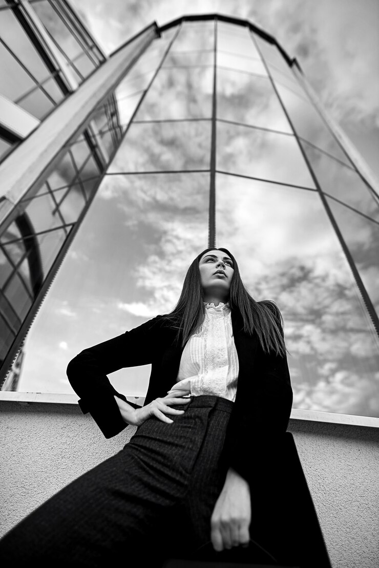 Mujer hermosa joven que se coloca delante de un edificio de oficinas de cristal grande.