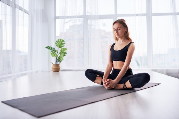 Mujer hermosa joven que hace yoga en una habitación moderna y luminosa con grandes ventanales. Concepto de yoga en casa.