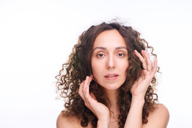 Mujer hermosa joven que mantiene las manos por la cara y el pelo ondulado largo oscuro mientras está de pie en blanco