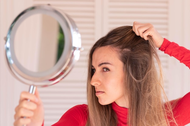 Mujer hermosa joven que mira al espejo que se ocupa de la alopecia debido a los tratamientos de blanqueamiento