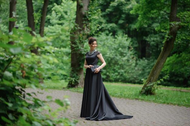 Mujer hermosa joven que presenta en un vestido negro en un parque. Sosteniendo un bolso en sus manos.