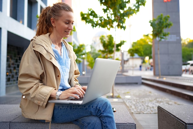 Mujer hermosa joven que trabaja en una computadora portátil sentada en el banco en la calle