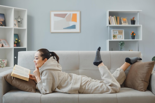 Mujer hermosa joven en ropa de casa recostada en un sofá en el interior de casa leyendo un libro feliz y positiva sonriendo ampliamente