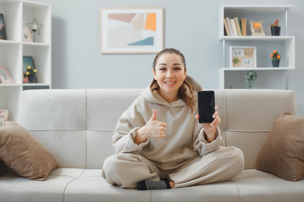 Mujer hermosa joven en ropa de casa sentada en un sofá en el interior de la casa sosteniendo un teléfono inteligente mostrando el pulgar hacia arriba sonriendo feliz y positivo