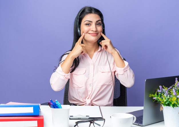 Mujer hermosa joven en ropa casual con auriculares con micrófono apuntando a su sonrisa sentado en la mesa con el portátil sobre fondo azul trabajando en la oficina