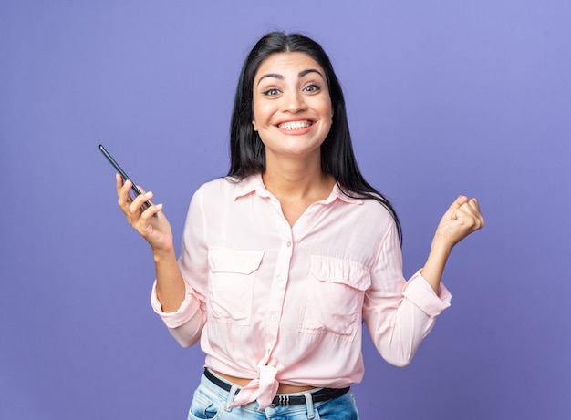 Mujer hermosa joven en ropa casual con smartphone feliz y emocionado apretando el puño de pie sobre la pared azul