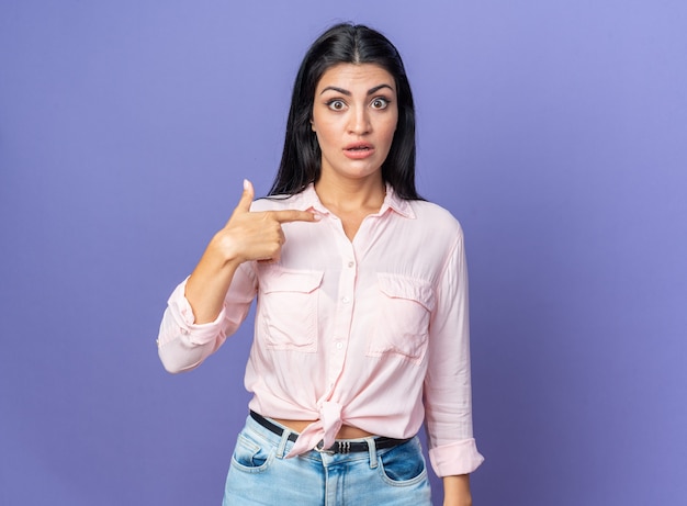 Mujer hermosa joven en ropa casual sorprendida señalando a sí misma de pie en azul