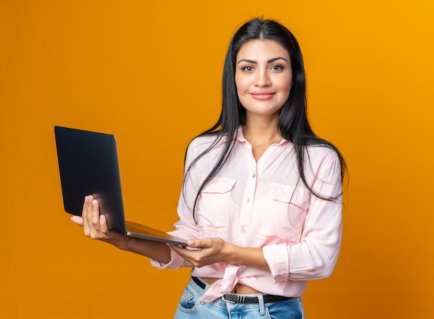 Foto mujer hermosa joven en ropa casual sosteniendo portátil feliz y positivo mirando al frente sonriendo confiado de pie sobre la pared naranja