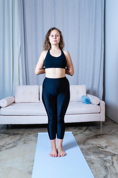 Mujer hermosa joven saludable en top deportivo y polainas practicando yoga en casa de pie en pose en estera de yoga