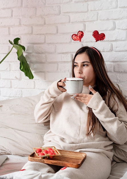 Mujer hermosa joven sentada en la cama celebrando el día de san valentín comiendo dulces y tomando café