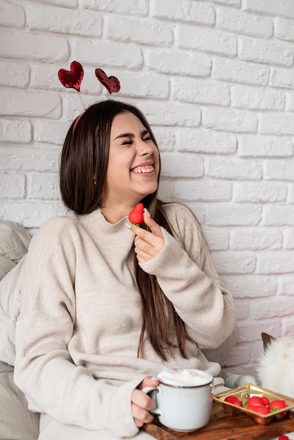 Mujer hermosa joven sentada en la cama con su perro celebrando el día de san valentín comiendo dulces y tomando café