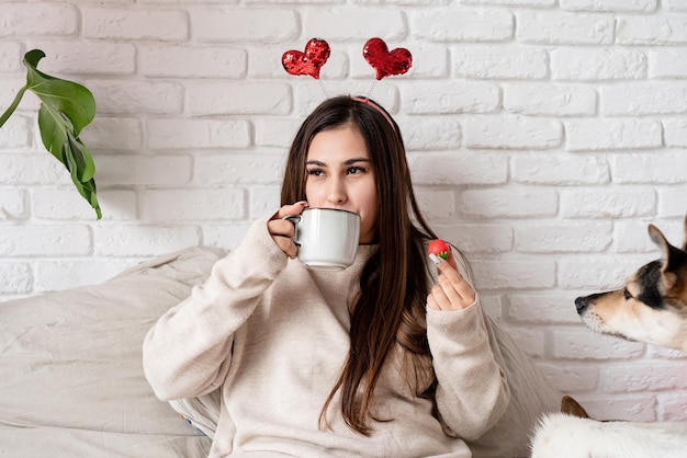 Mujer hermosa joven sentada en la cama con su perro celebrando el día de san valentín comiendo dulces y tomando café