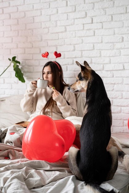 Mujer hermosa joven sentada en la cama con su perro celebrando el día de san valentín comiendo dulces y tomando café