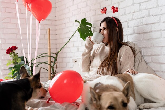 Mujer hermosa joven sentada en la cama con su perro celebrando el día de san valentín comiendo dulces y tomando café