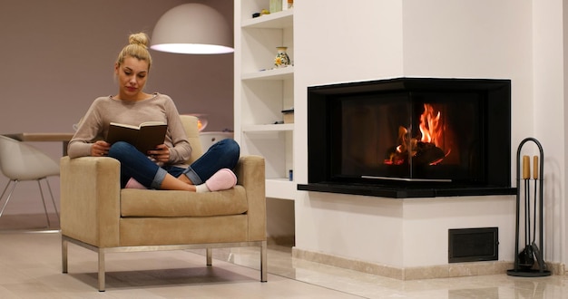 Mujer hermosa joven sentada frente a la chimenea en casa en un frío día de otoño y leyendo un libro