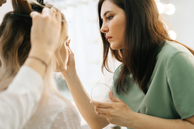 Mujer hermosa joven sentada en una silla frente al espejo y obtiene un maquillaje profesional en el salón Trabajos de maquilladores profesionales Concepto de trabajo entre bastidores