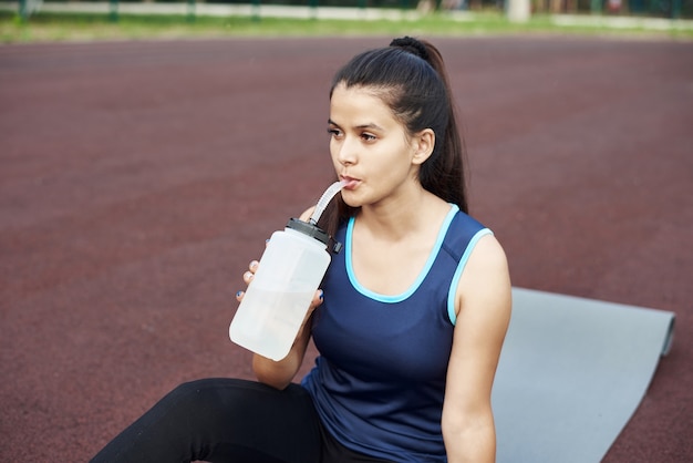 Mujer hermosa joven se sienta en una estera de yoga al aire libre y bebe agua de una botella de agua reutilizable. Deportes al aire libre.