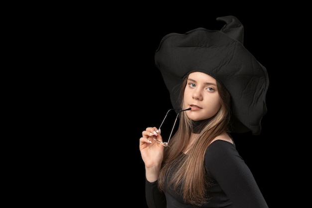 Mujer hermosa joven con sombrero de bruja puntiagudo con gafas en las manos Disfraz de Halloween Fondo negro Copiar espacio