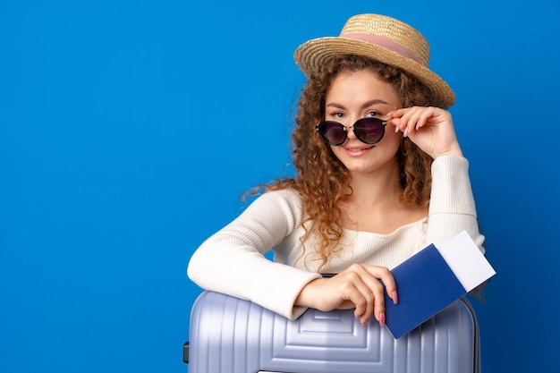 Mujer hermosa joven con sombrero de vacaciones con maleta contra el fondo azul.