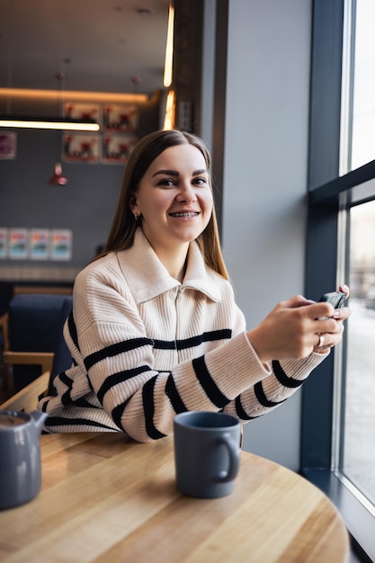 Mujer hermosa joven sosteniendo y mirando el teléfono inteligente mientras está sentado en la cafetería. Estudiante universitario feliz usando teléfono móvil. La empresaria bebe café y sonríe.