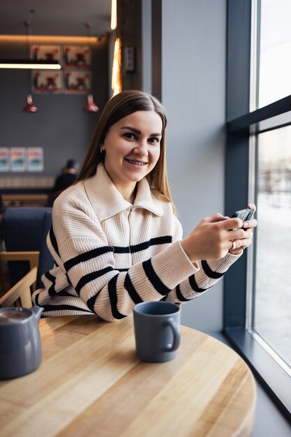 Mujer hermosa joven sosteniendo y mirando el teléfono inteligente mientras está sentado en la cafetería Estudiante universitario feliz usando teléfono móvil Empresaria bebe café y sonríe