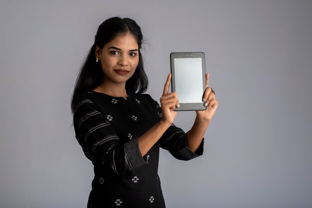 Mujer hermosa joven sosteniendo y mostrando la pantalla en blanco del teléfono inteligente o móvil o tableta en un gris.