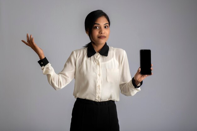 Mujer hermosa joven sosteniendo y mostrando smartphone con pantalla en blanco o teléfono móvil o tableta en una pared gris.
