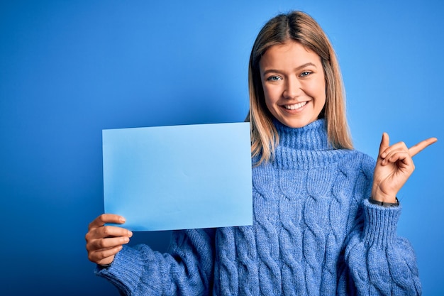 Mujer hermosa joven sosteniendo una pancarta de pie sobre un fondo azul aislado muy feliz señalando con la mano y el dedo a un lado