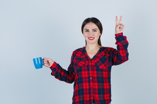 Mujer hermosa joven sosteniendo la taza mientras muestra un gesto de paz en camisa casual, jeans y mirando dichoso, vista frontal.