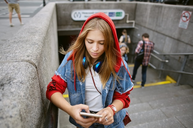 Mujer hermosa joven en sudadera con capucha roja está de pie en la entrada del metro con el teléfono móvil en las manos.