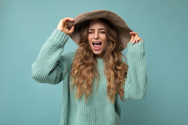 Mujer hermosa joven con suéter azul. La mujer positiva muestra emociones faciales sinceras. Modelo divertido aislado en la pared azul con espacio vacío.